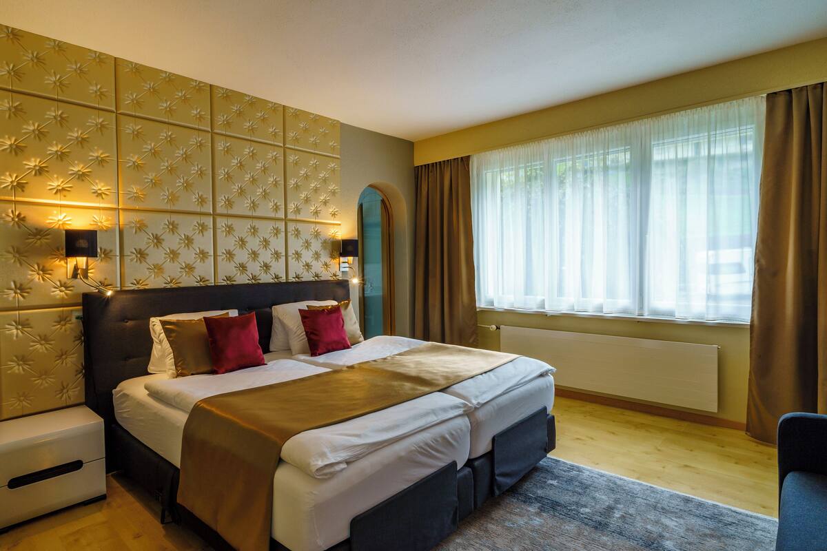 A hotel room with a large bed, a headboard with a floral pattern, a nightstand, and a window with curtains.