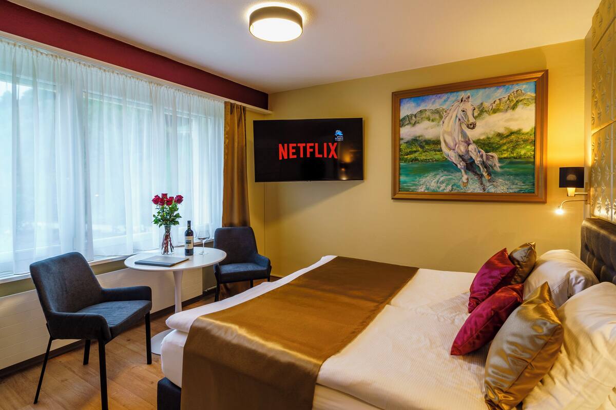 A hotel room with a bed, a desk, a chair, a TV displaying 'NETFLIX', and a painting on the wall.