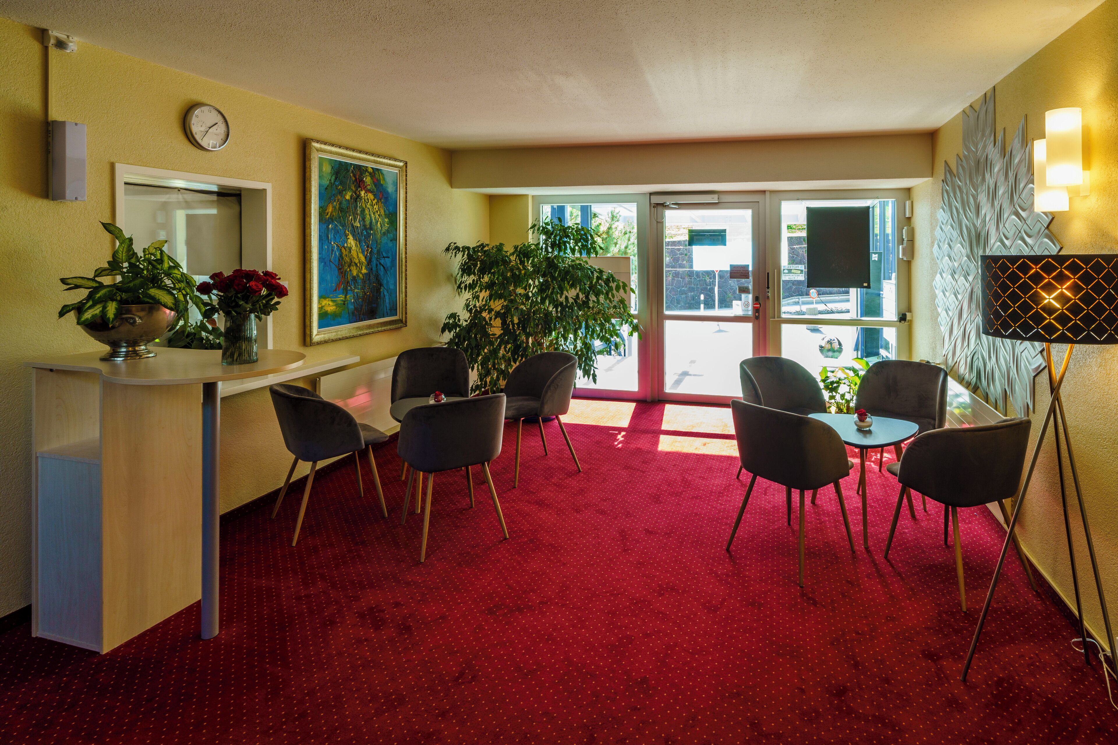Habitación de hotel con alfombra roja, una zona de comedor con sillas, una planta grande en maceta y un reloj en la pared.