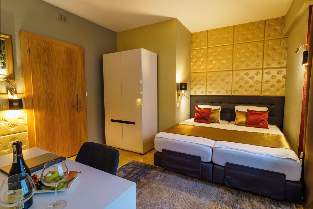 A hotel room with a bed, a desk with a laptop, a wardrobe, and a dining table with fruits and wine.