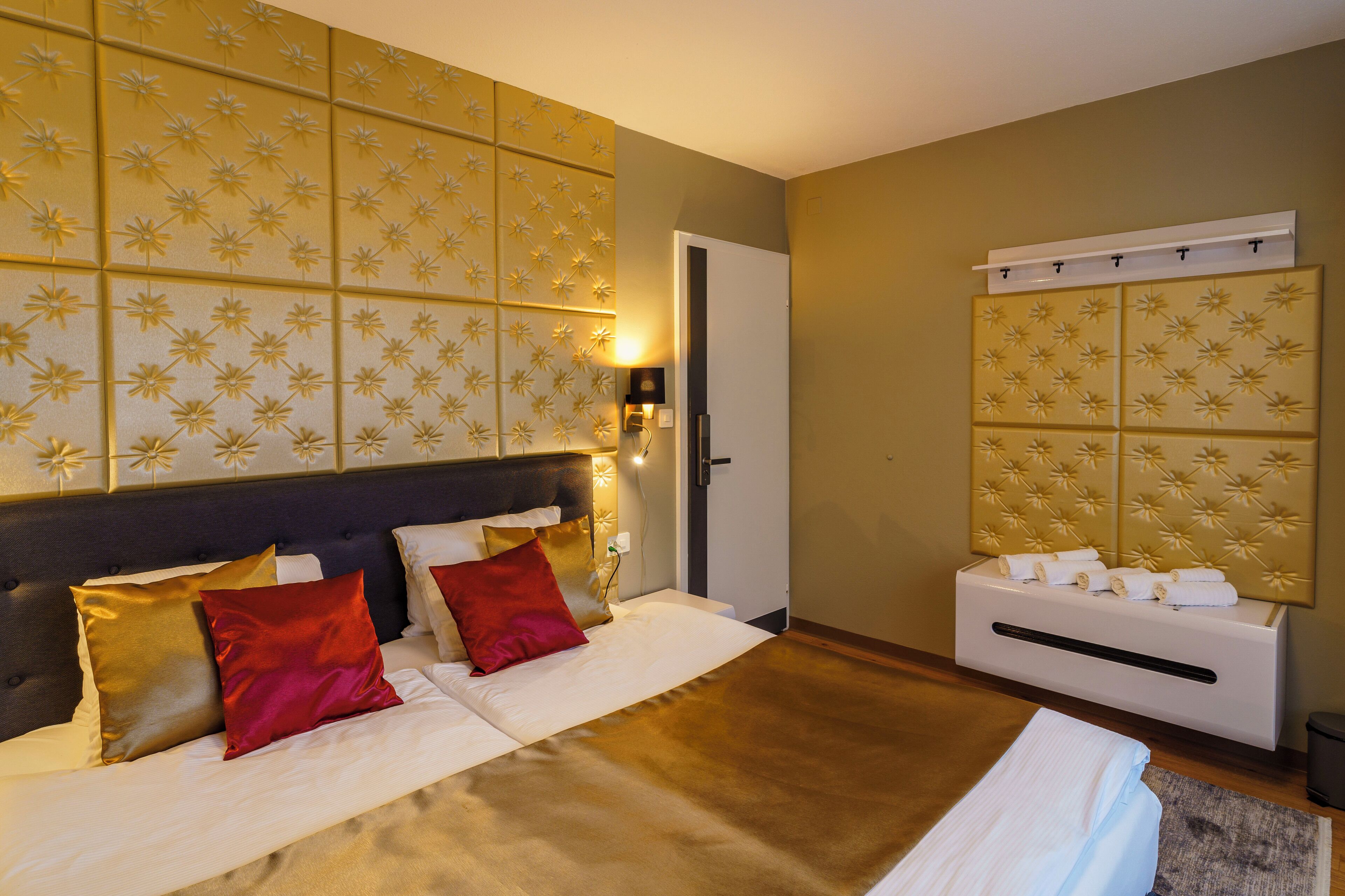 Una habitación de hotel con una cama grande, un cabecero con una lámpara, un tocador adosado a la pared con toallas y una puerta a la derecha.