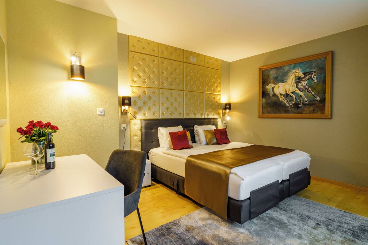 A hotel room with a bed, bedside lamps, a desk with a vase of flowers, and a painting on the wall.