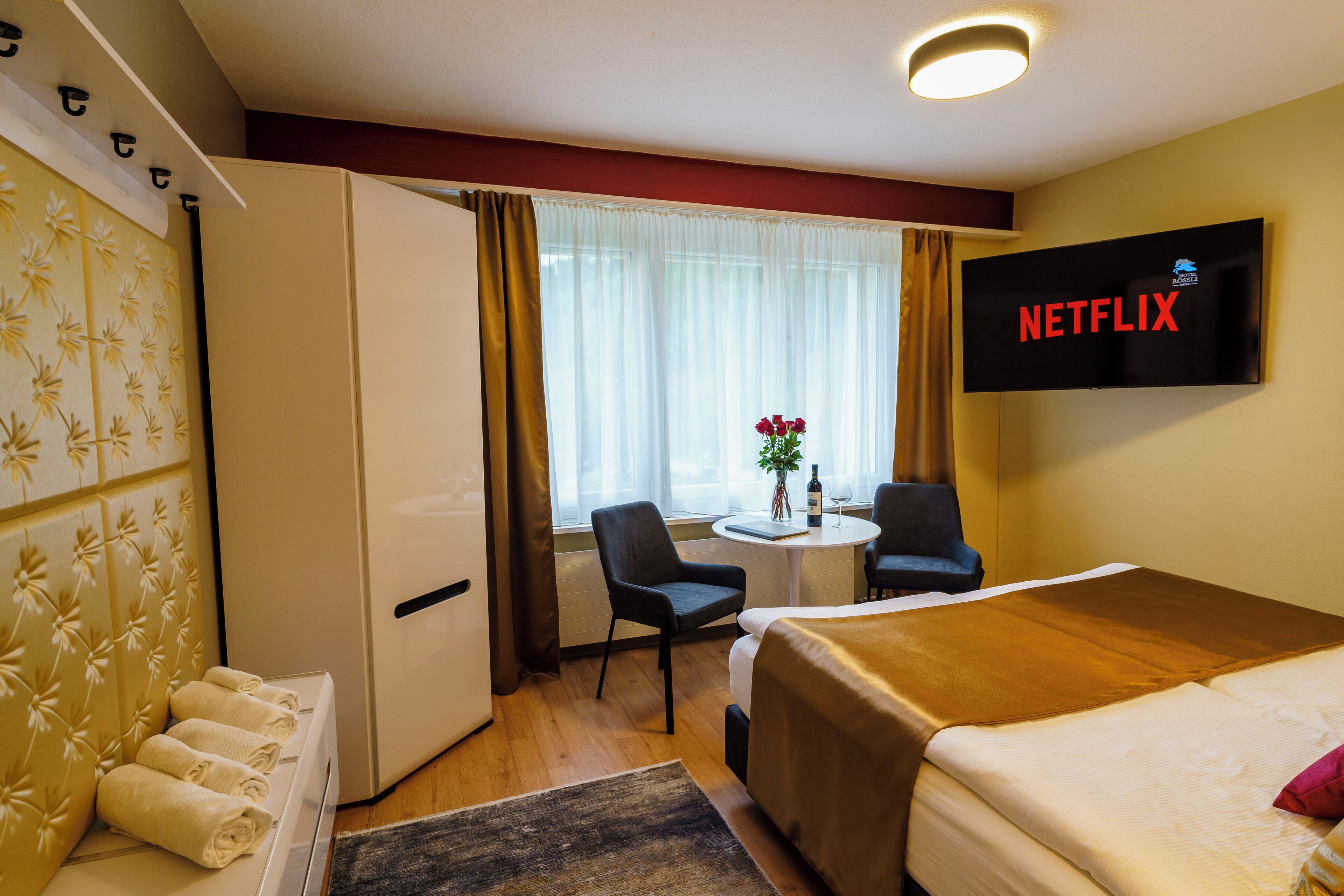 A hotel room with a bed, a desk with a chair, a TV displaying 'NETFLIX', and a window with curtains.
