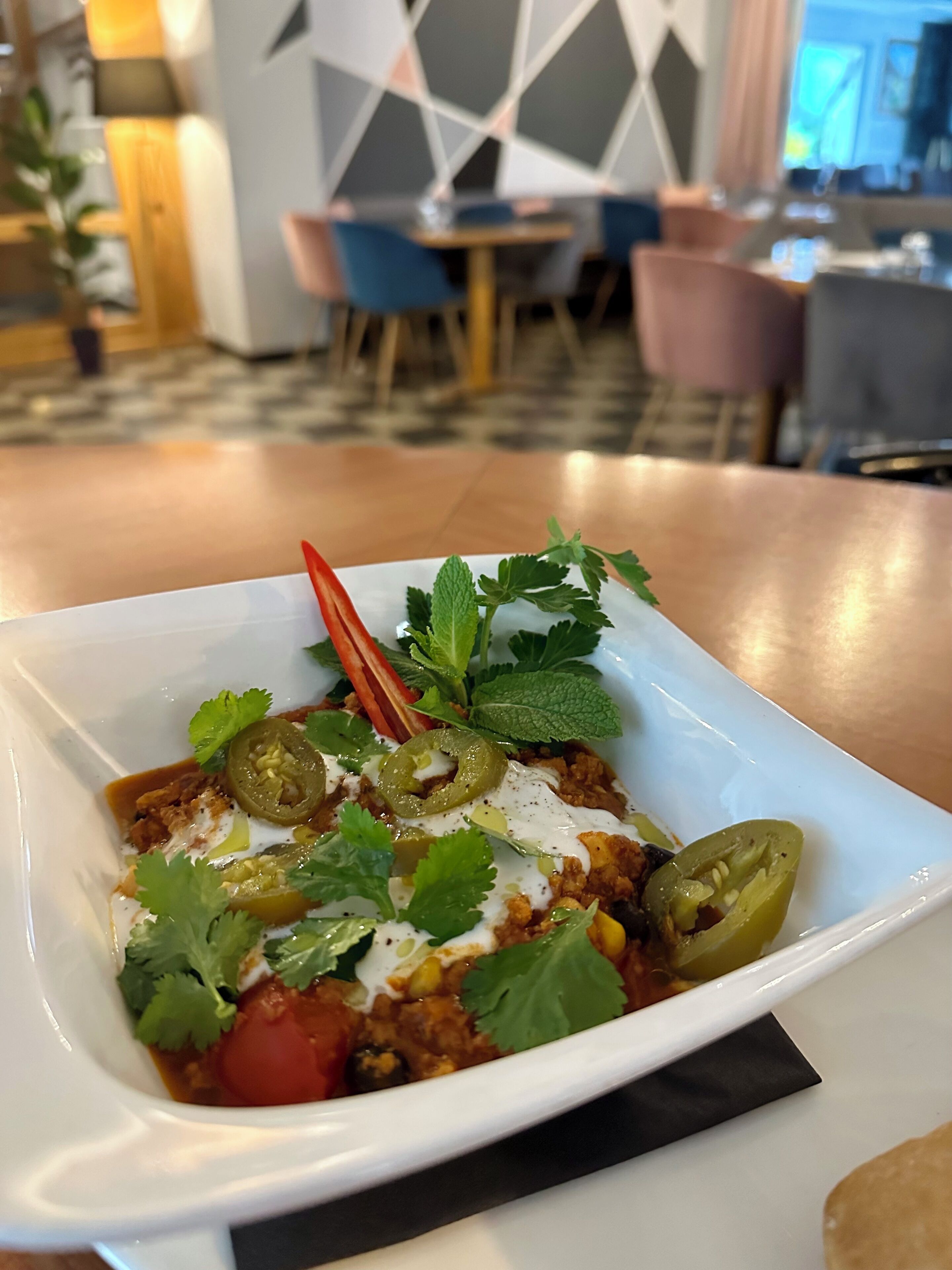Ein weißer Teller mit einer Speise, garniert mit Koriander, Jalapeños und einer roten Chili, auf einem Tisch in einem Restaurant mit geometrischer Wandgestaltung und modernen Möbeln.