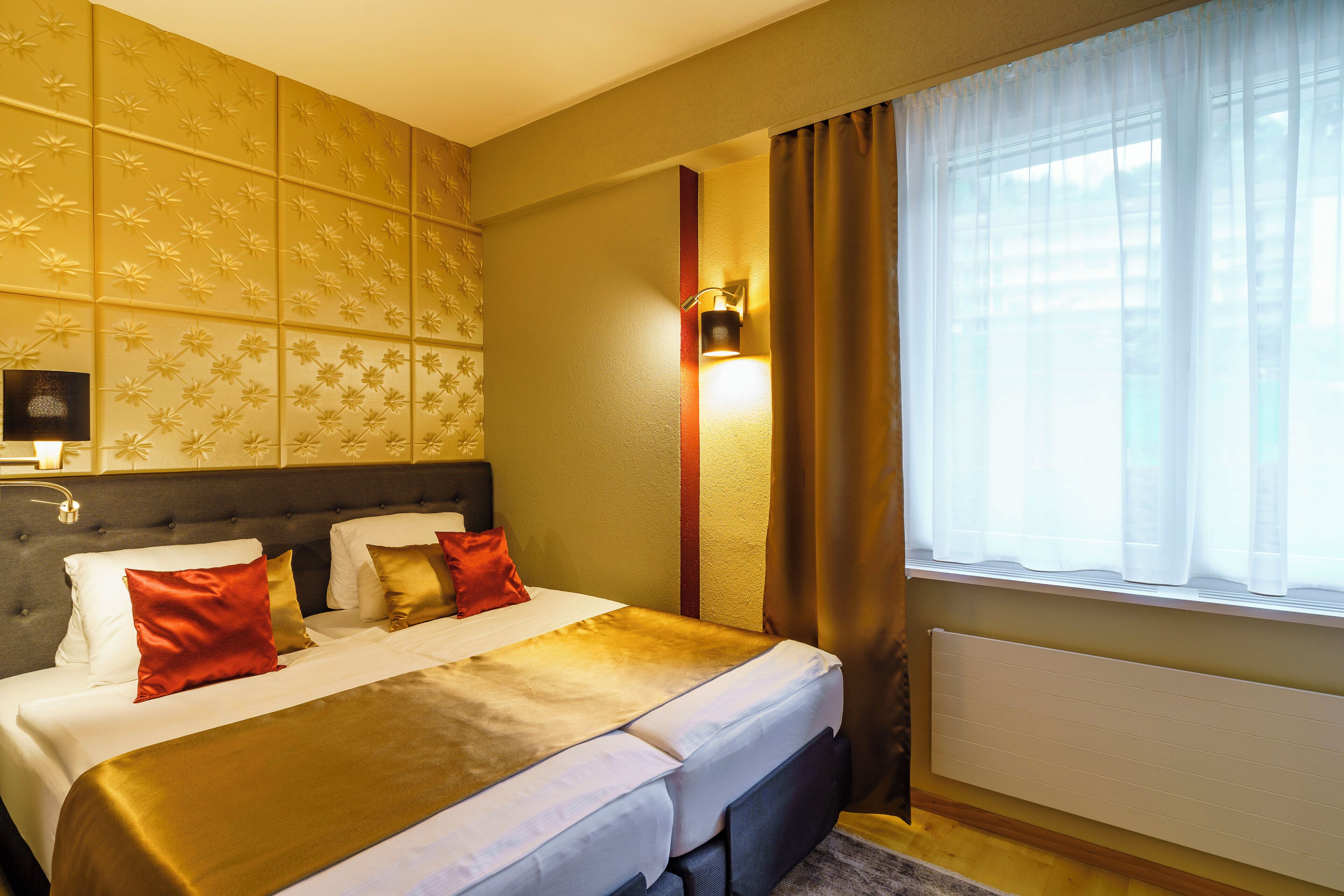 Ein Hotelzimmer mit einem großen Bett, einem Kopfteil, einem Nachttisch, einer Lampe und einem Fenster mit Vorhängen.