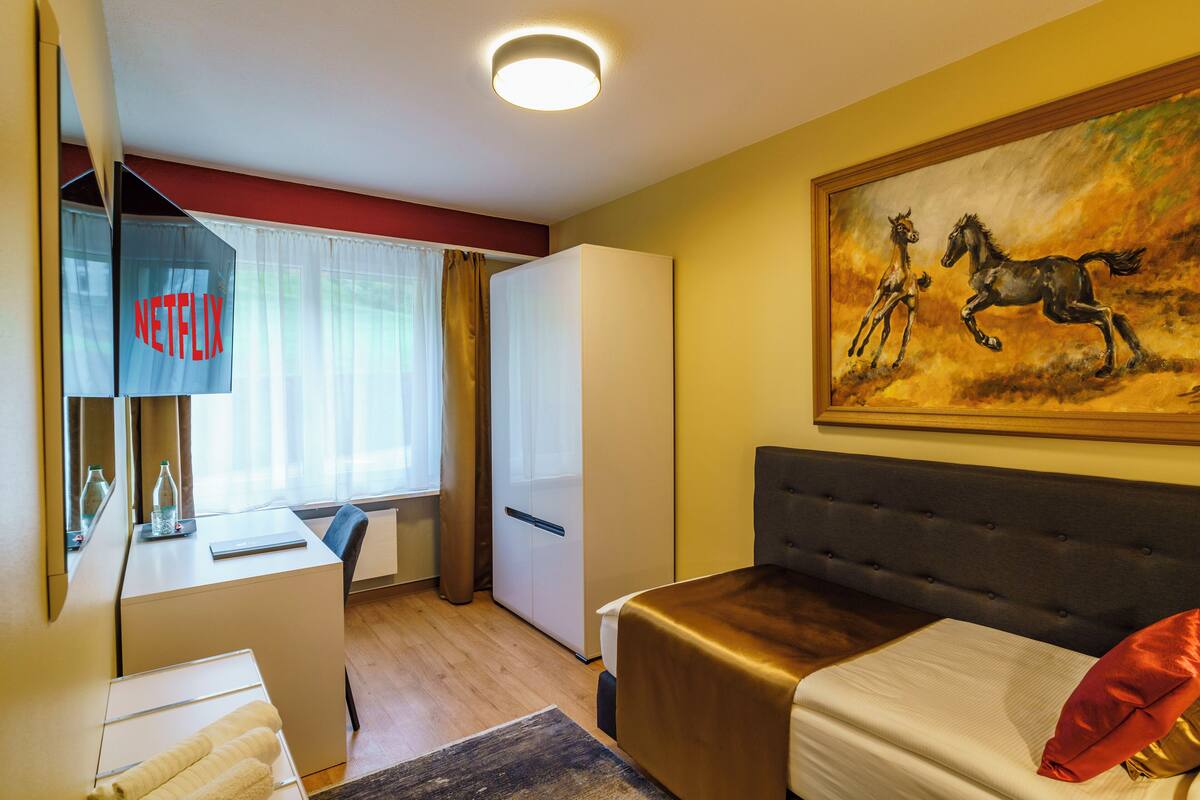 A hotel room with a bed, a desk, a wardrobe, and a painting of horses.