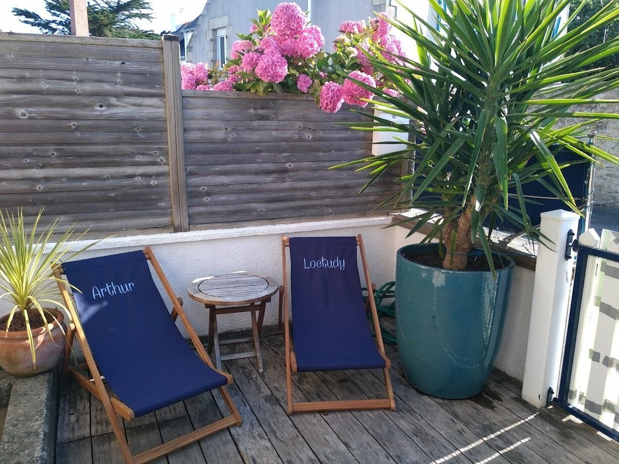 Zwei Lounge-Stühle mit den Aufschriften 'Arthur' und 'Loctudy', ein Blumentopf und ein Holzzaun mit rosa Blüten.