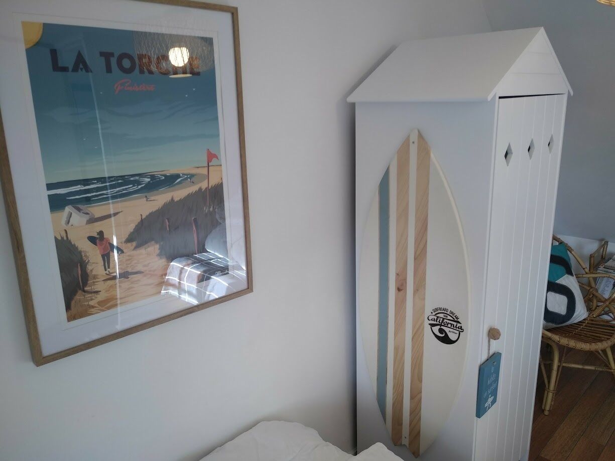 A room with a framed poster of a beach scene, a surfboard leaning against a cabinet, and a wicker chair.