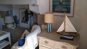 Un salon chaleureux avec une petite table d’appoint en osier, une lampe, un tableau encadré représentant un phare et une maquette de petit voilier.