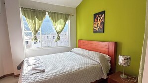 Une chambre à coucher avec un lit, une table de chevet avec une lampe, une fenêtre avec des rideaux et un paillasson.