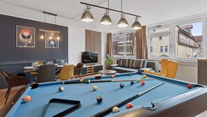 Ein Billardtisch mit Kugeln, ein Fernseher und Sitzbereiche in einem Raum mit großen Fenstern und Blick auf die Stadt.