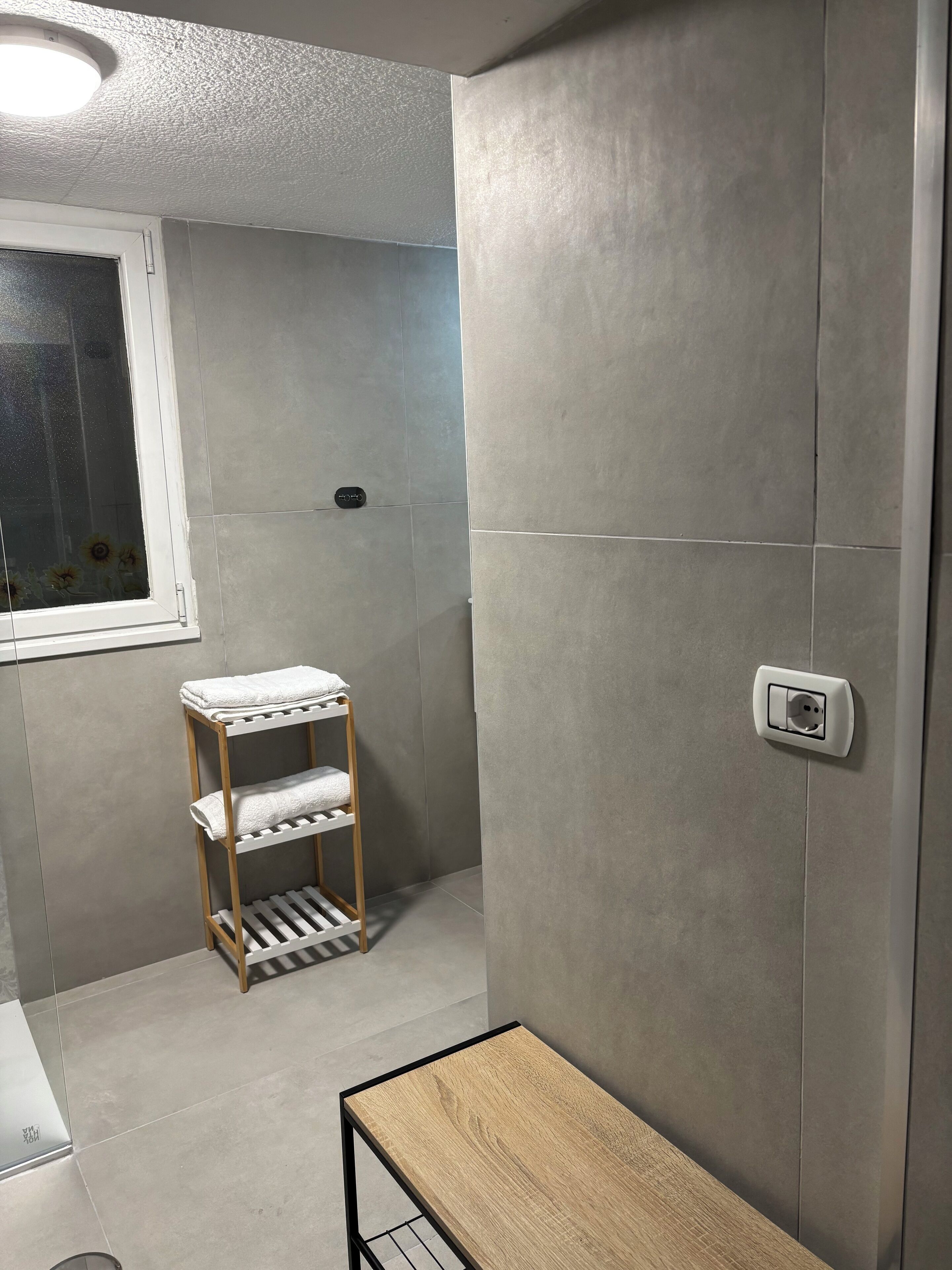 A bathroom with grey tiles, a wooden towel rack, a small table, and a window with frosted glass.