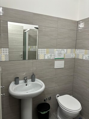 Une salle de bain équipée d’un lavabo, d’un miroir, de toilettes et d’une poubelle.