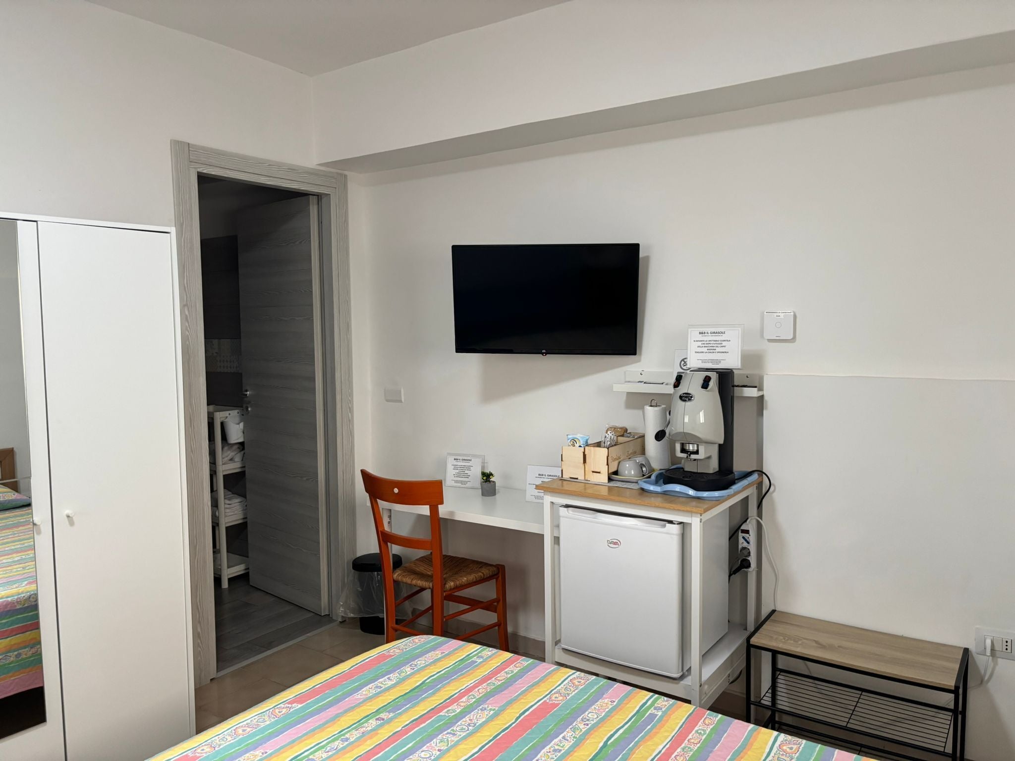 Um quarto pequeno com uma cama, uma escrivaninha com cafeteira, uma televisão e uma cadeira.