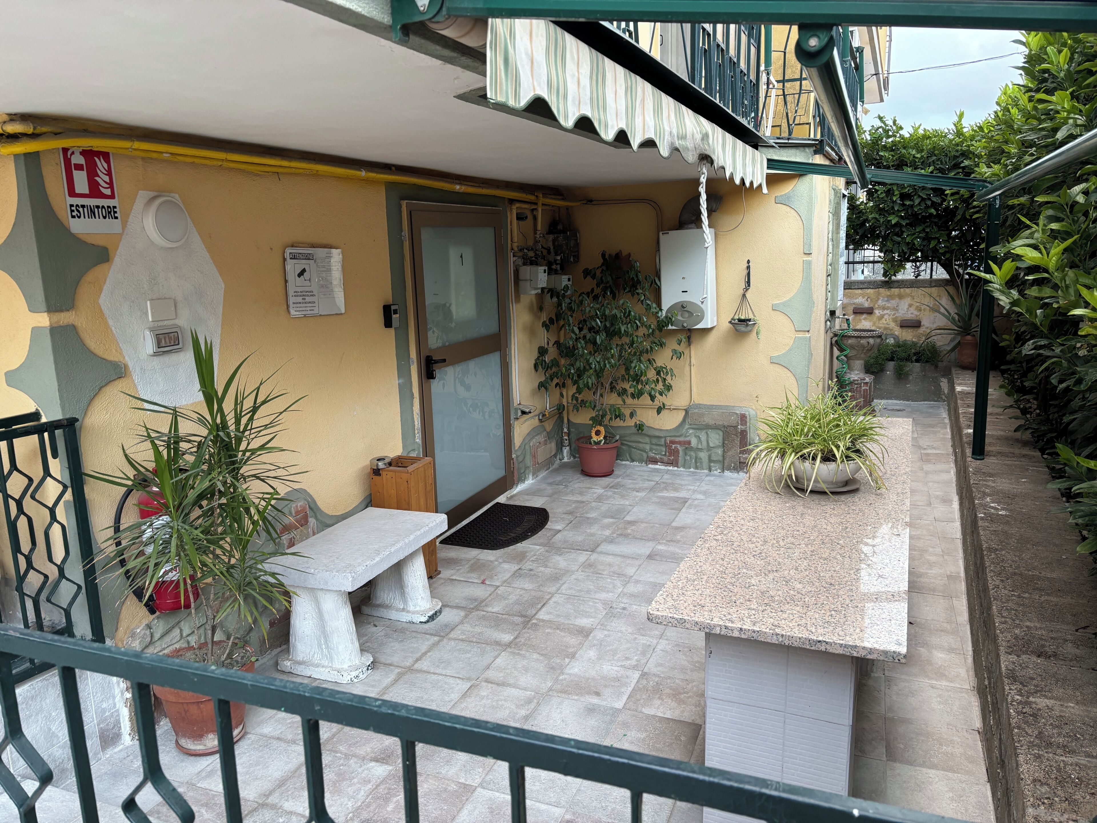 A small outdoor area with a bench, potted plants, a door, and a fire extinguisher sign.