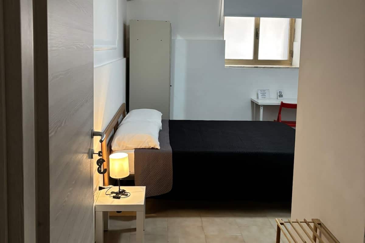 A small, compact room with a bed, a bedside table with a lamp, a wardrobe, a window with blinds, and a small desk with a chair.
