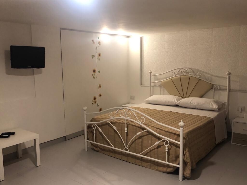 A bedroom with a white metal bed frame, a bedside table, a flat-screen TV mounted on the wall, and a door with a floral curtain.