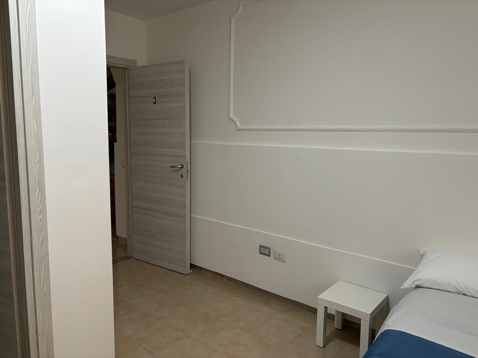 Un dormitorio con una cama, una mesita de noche pequeña, un armario blanco y una puerta.