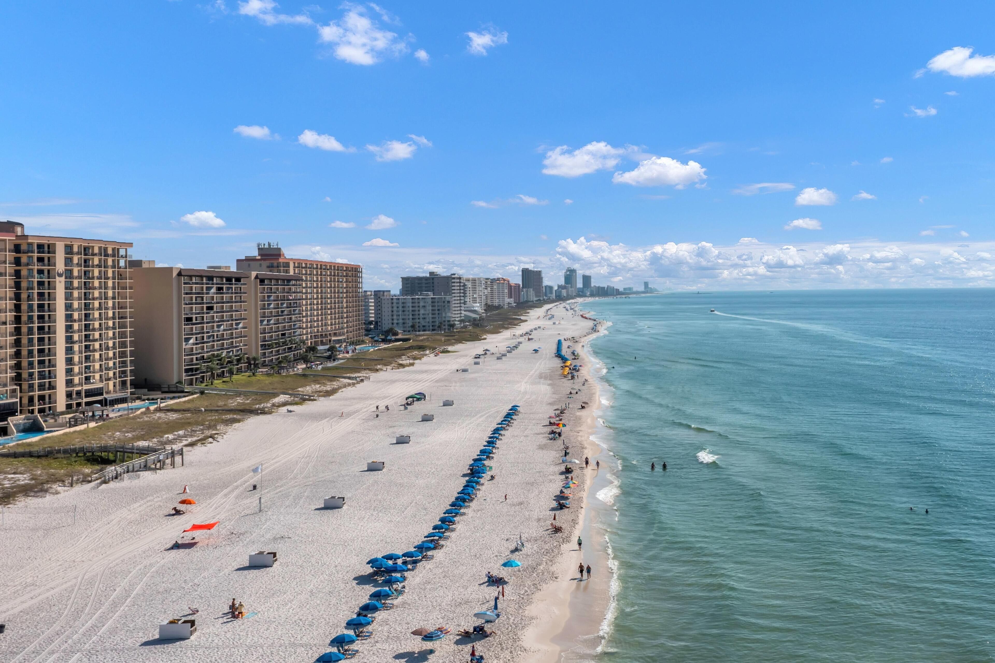 Une plage bordée de rangées de parasols, un rivage sablonneux et des bâtiments le long de la côte.