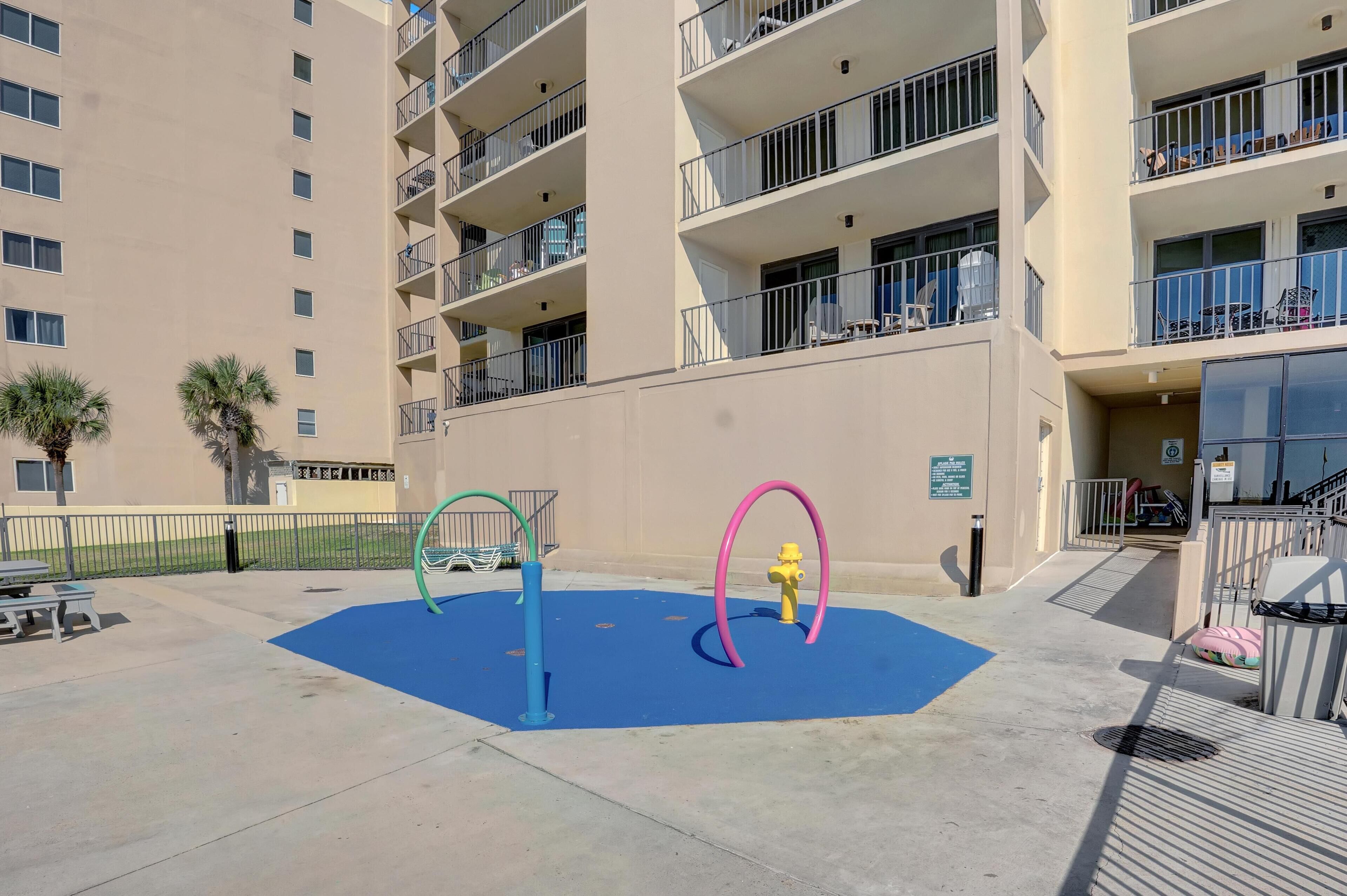 Un parque infantil con juegos de colores, un tapete azul y un edificio con balcones al fondo.