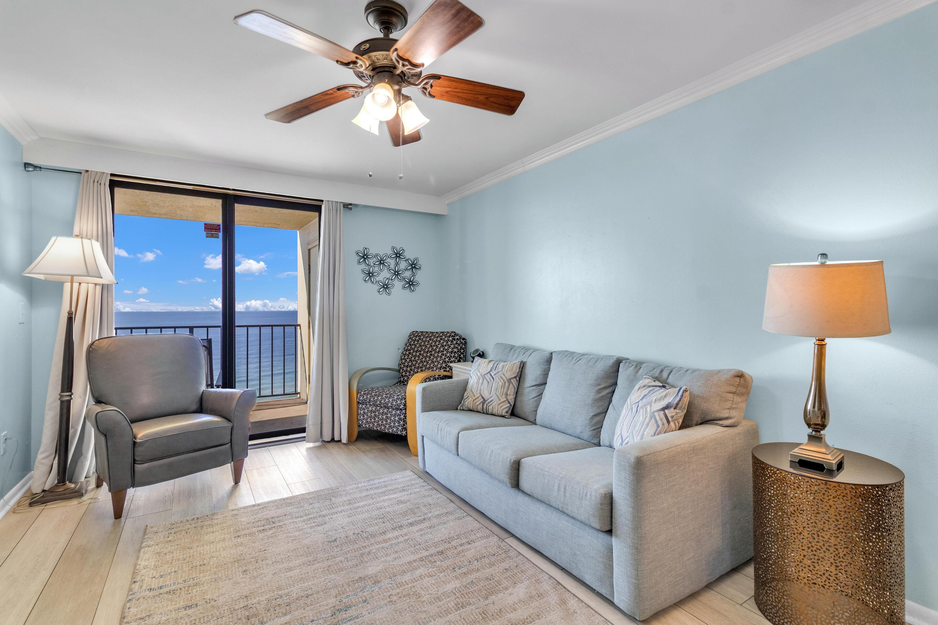 A living room with a ceiling fan, a sofa, a chair, a lamp, and a view of the ocean through a sliding door.