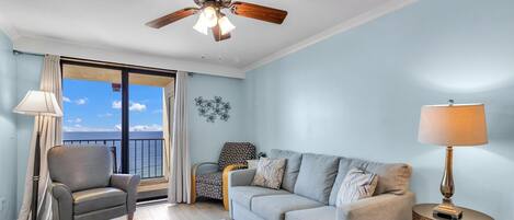 A living room with a ceiling fan, a sofa, a chair, a lamp, and a view of the ocean through a sliding door.