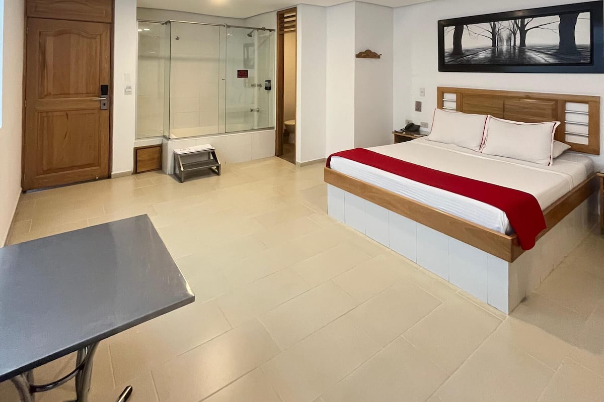 A modern hotel room with a bed, a desk, a glass shower enclosure, and a painting on the wall.