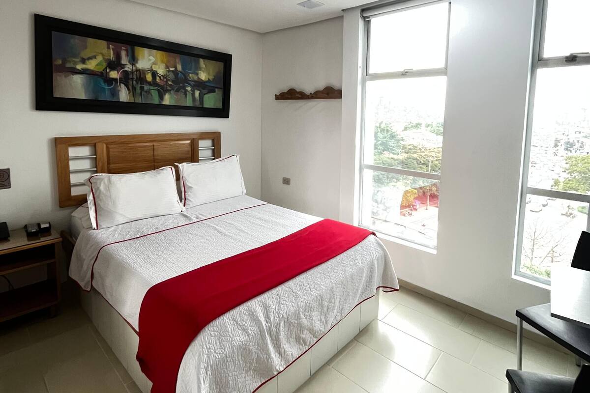 A bedroom with a bed, a desk, a chair, a painting, and a window.