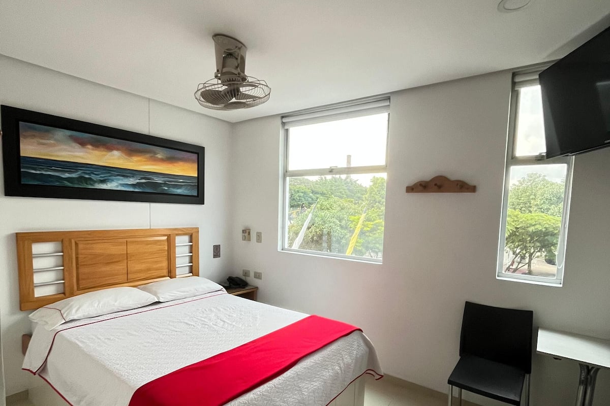 A bedroom with a bed, a desk, a chair, a TV, and a window with a view of greenery.