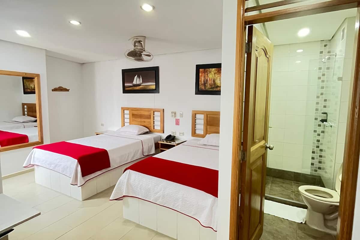 A hotel room with two beds, a desk, a mirror, a ceiling fan, and a bathroom visible through an open door.
