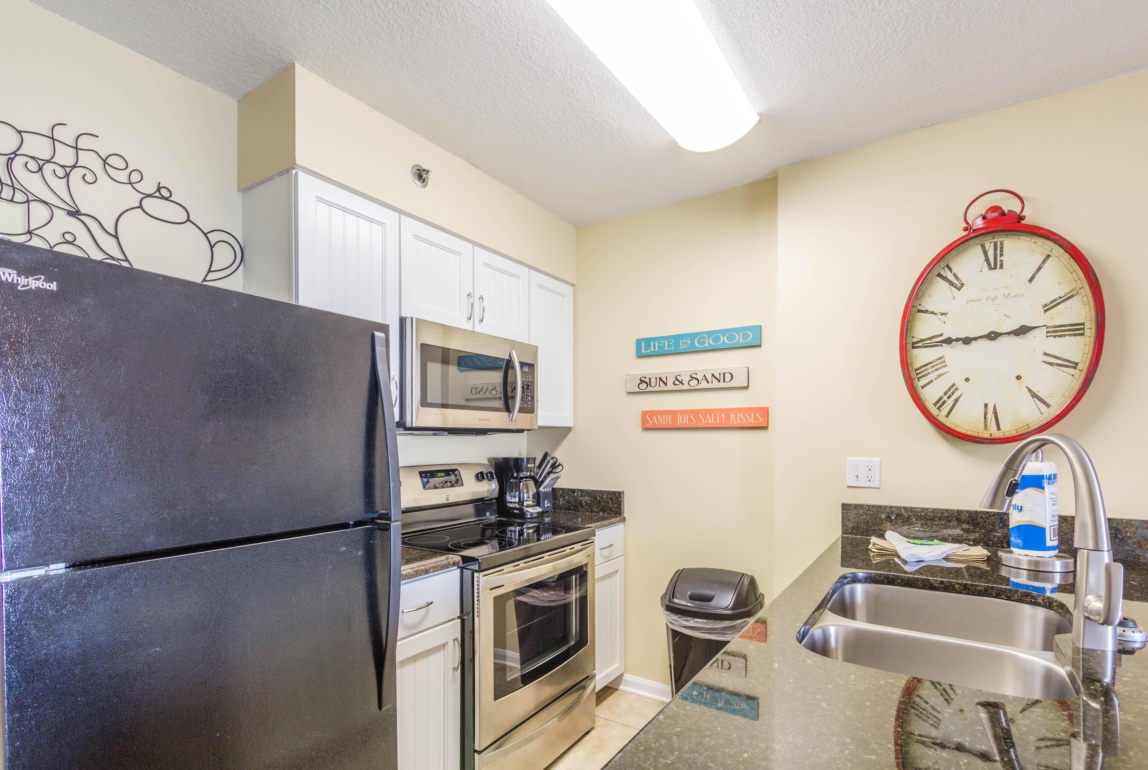 A kitchen with a refrigerator, microwave, oven, sink, and a clock on the wall.