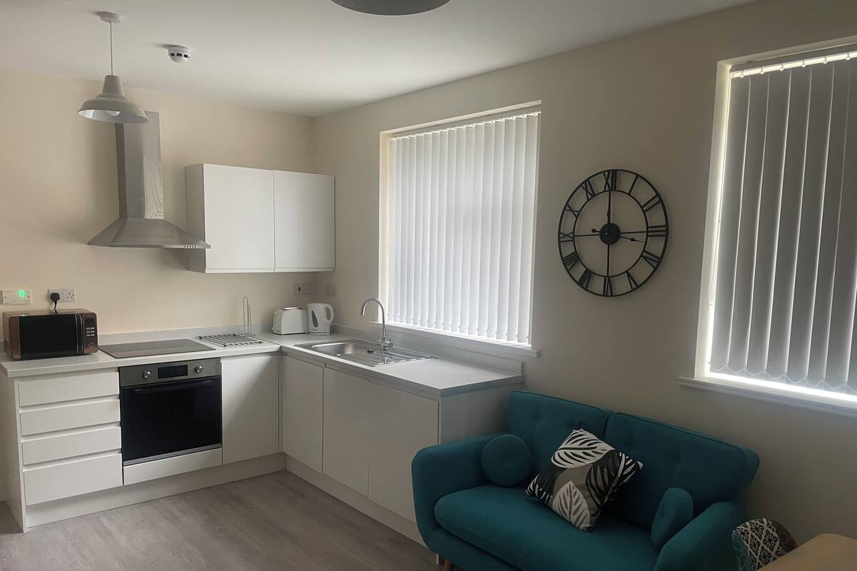 A compact kitchen with a microwave, toaster, and sink. There is a blue sofa with decorative pillows and a wall clock.