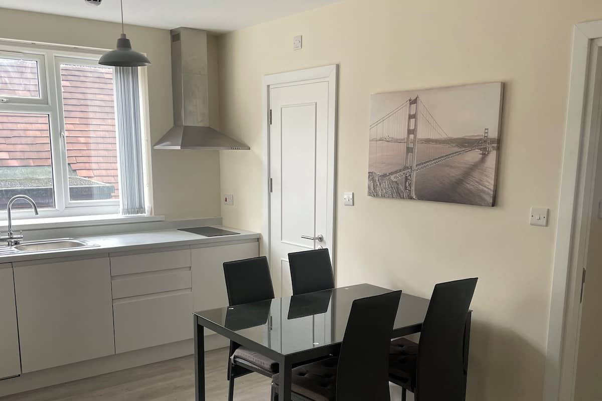 A modern kitchen with a dining area, featuring a glass table and black chairs, a white door, and a painting of a bridge.