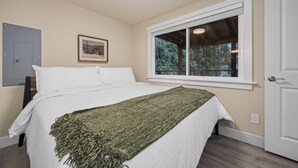 Un dormitorio con cama, ventana con vista a árboles, puerta y lámpara fijada a la pared.