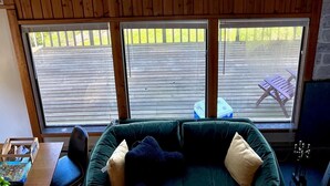 A cozy room with a wooden ceiling, a green sofa with cushions, and three large windows offering views of a lake, trees, and buildings. - Charming Pet-Friendly Cabin with Sunset Views Over Birch Bay (Blaine)