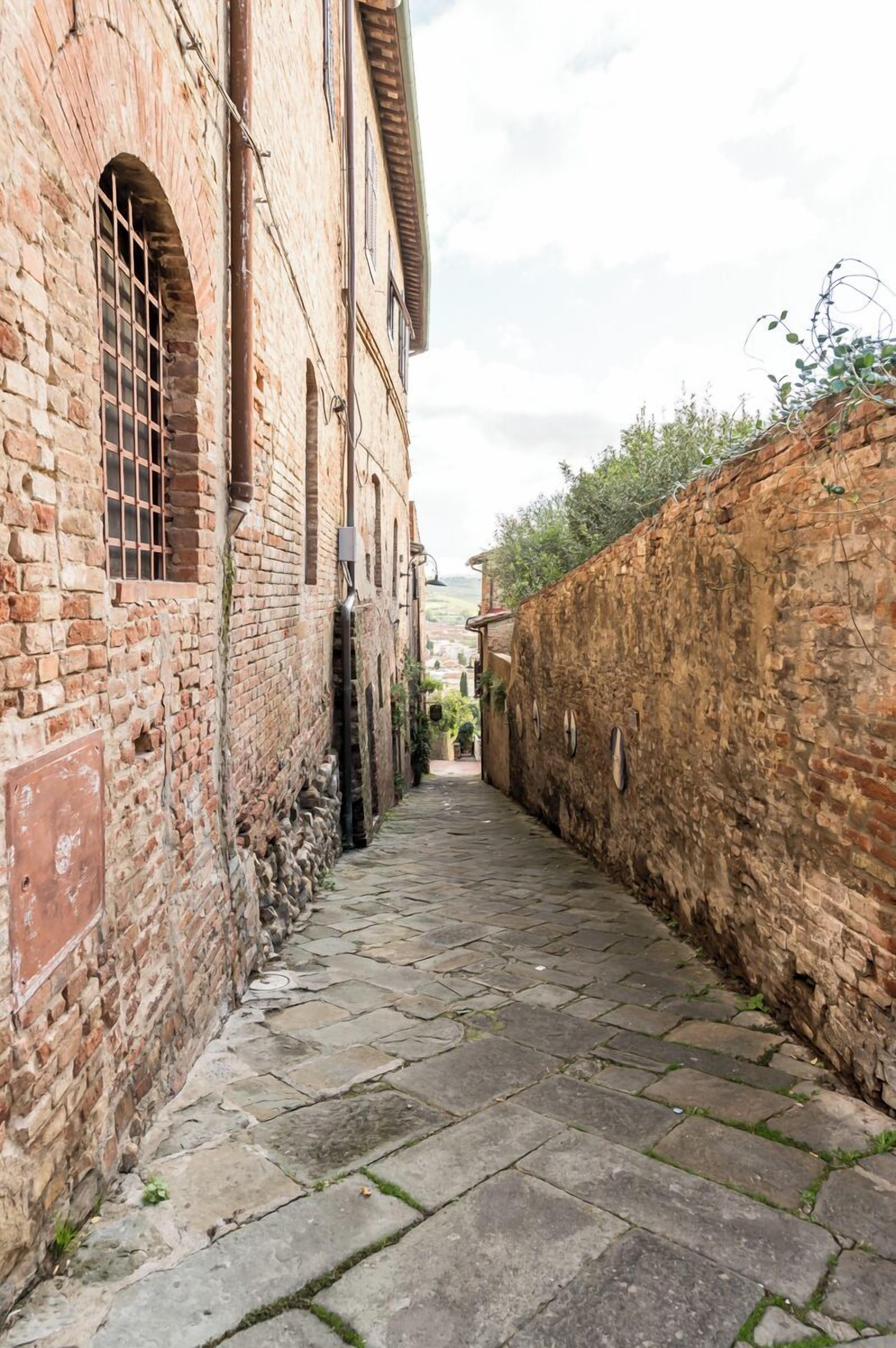 A narrow cobblestone alley flanked by brick buildings with arched windows and a stone wall on one side.