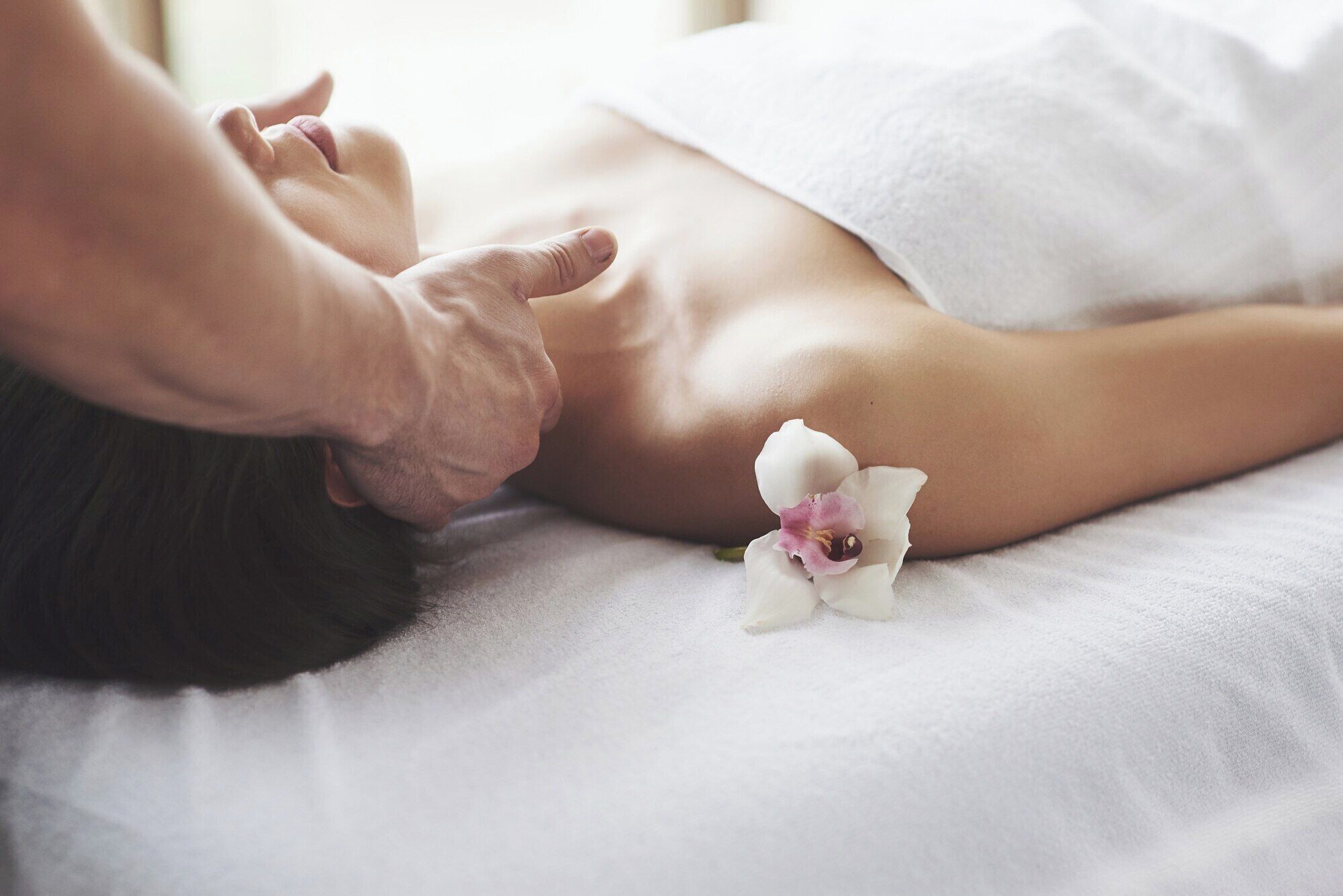Une personne reçoit un massage sur un lit blanc, avec une petite orchidée blanche posée à côté.