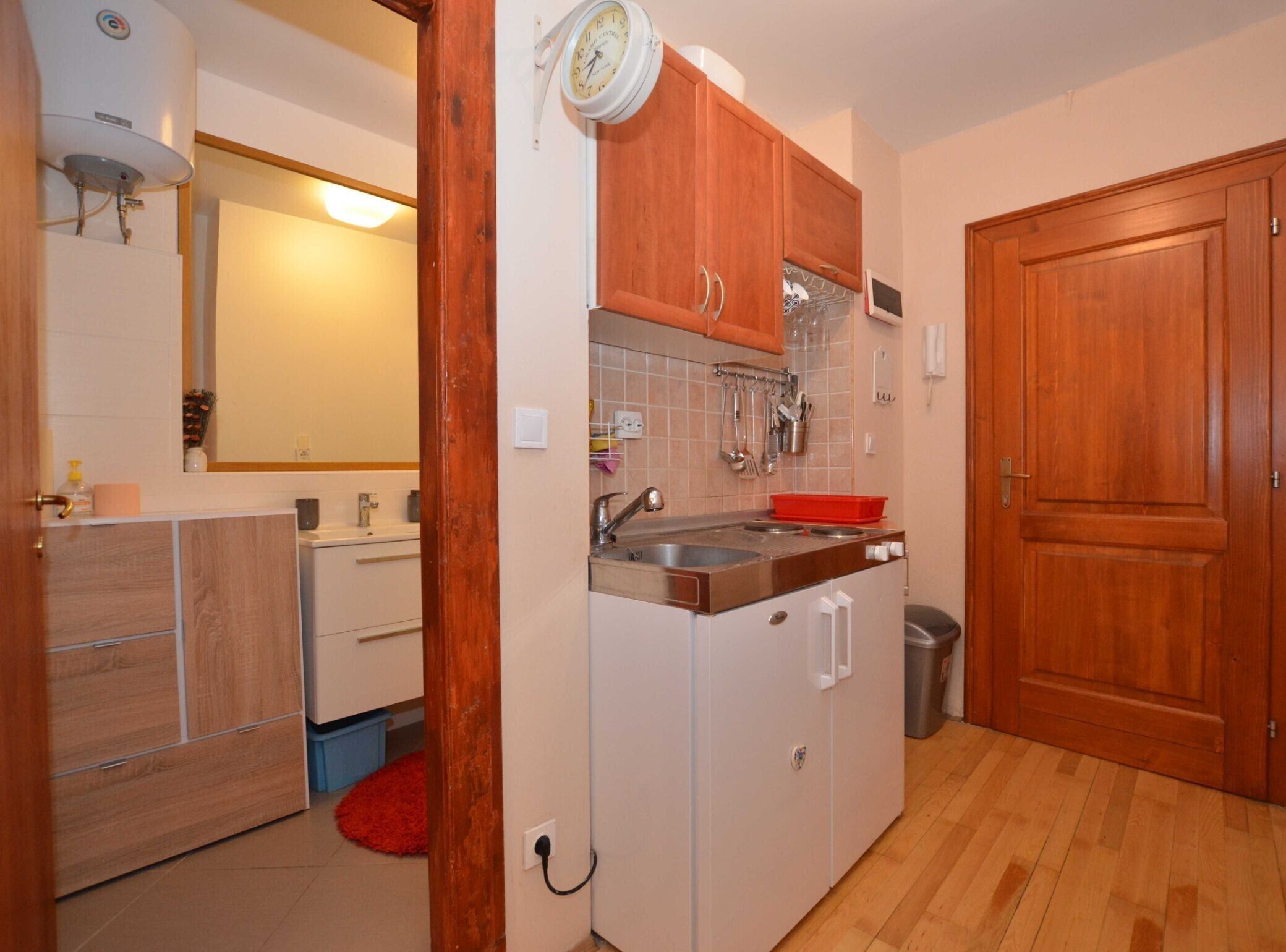 A compact kitchen with wooden cabinets, a sink, and a stove. There is a mirror above the sink and a clock on the wall.