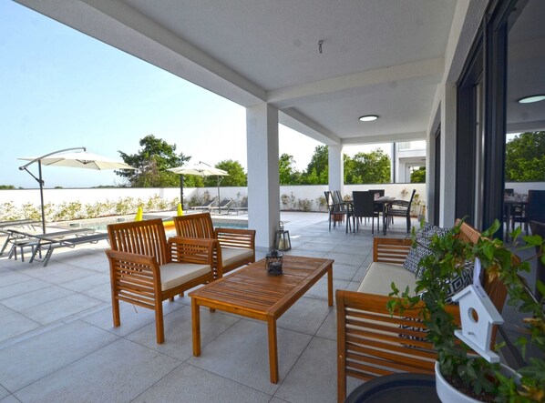 Relax by the pool table or enjoy a meal at the outdoor dining area, shaded by umbrellas and surrounded by lush greenery and a serene water view.