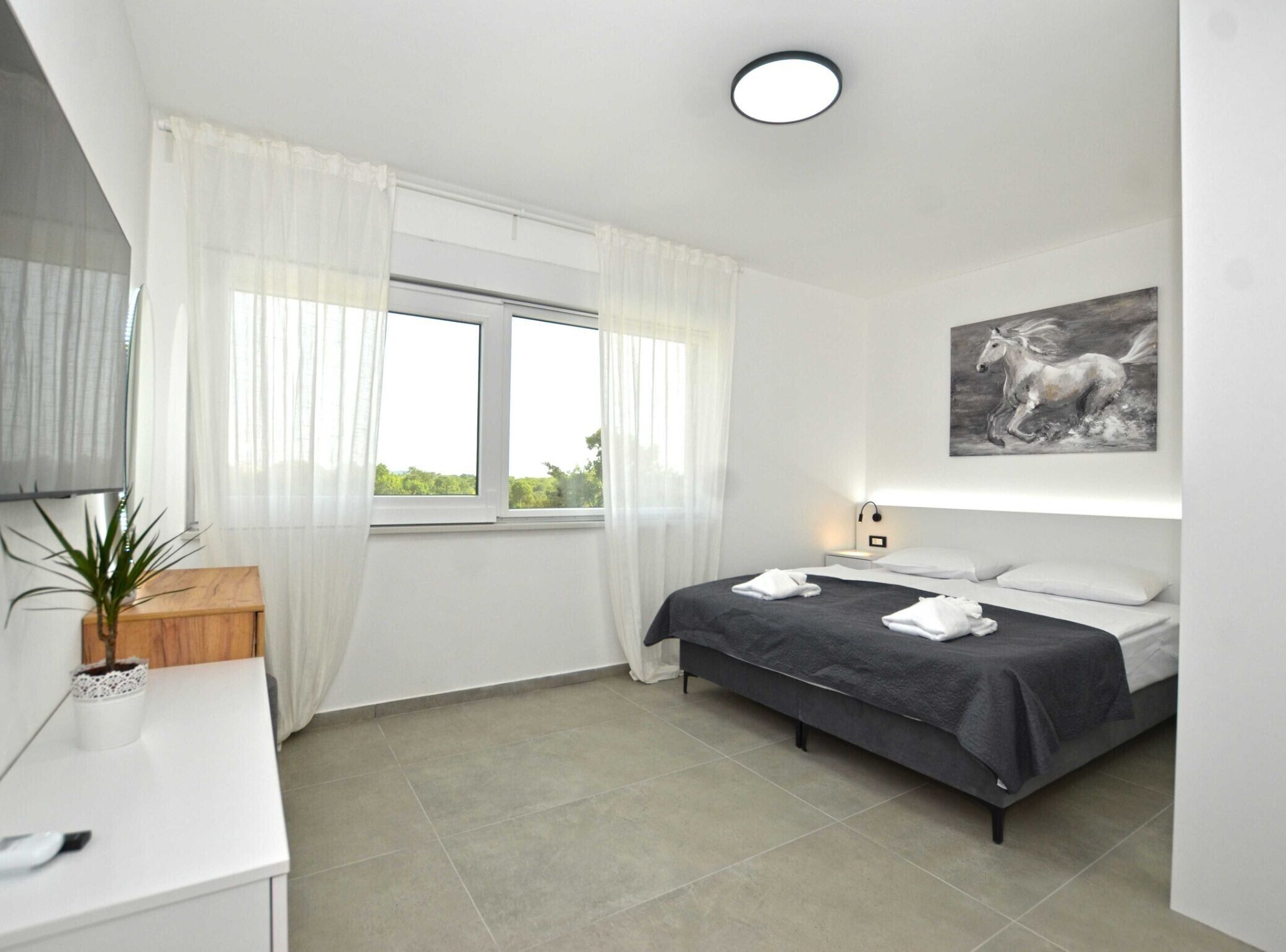 Un dormitorio con cama, mesita de noche, lámpara, ventana con cortinas, un cuadro en la pared y una planta en maceta.