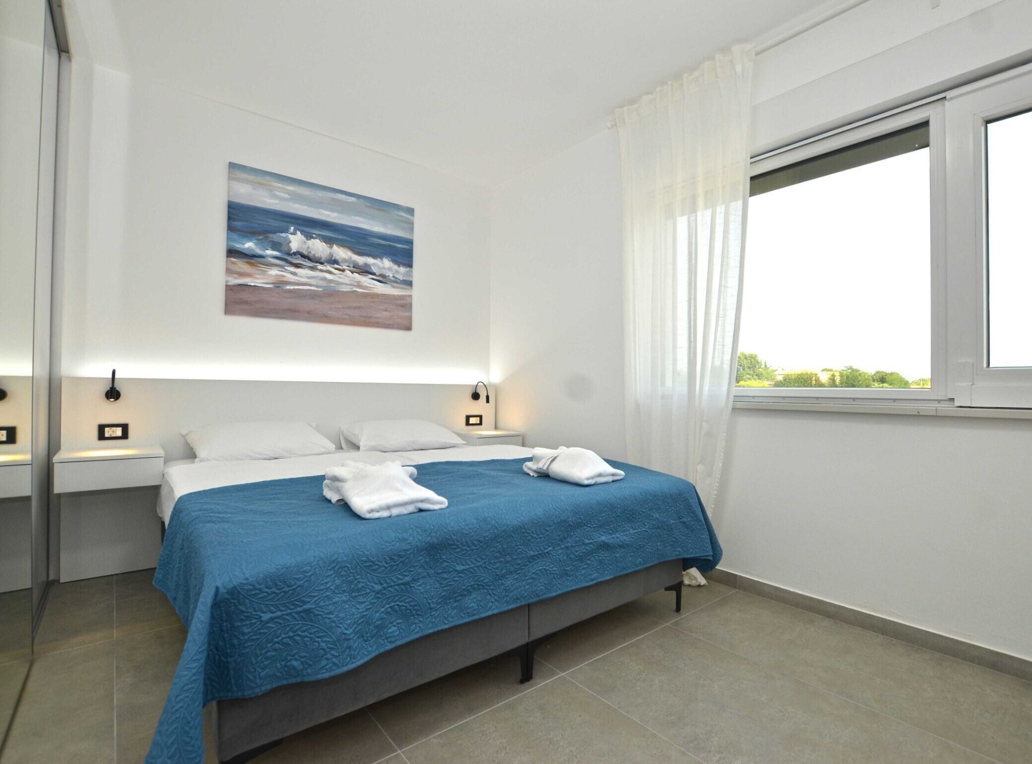 A modern hotel room with a large bed, white linens, a blue bedspread, and a painting of a seascape on the wall.