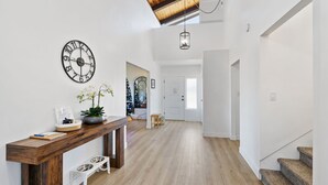 Un hall spacieux avec un plancher en bois, une grande horloge murale, une plante en pot et une petite table sur laquelle se trouve une gamelle pour chien.