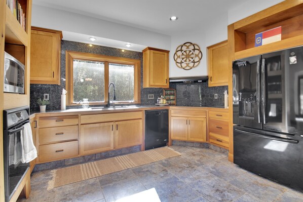 A modern kitchen with wooden cabinets, a black refrigerator, and a window with a view of greenery. - Big Sam's Riverside Retreat (Stanfield)