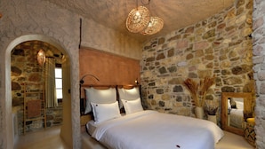A bedroom with a stone wall, a wooden headboard, a large bed with white linens, a hanging light fixture, and a mirror on the wall. - Chi House (Ayvalik)