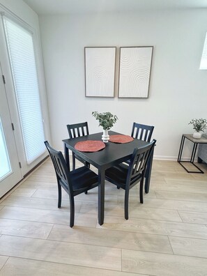 Un coin repas avec une table en bois foncé, quatre chaises, deux sets de table rouges, un vase blanc contenant une plante et deux œuvres d’art encadrées accrochées au mur.