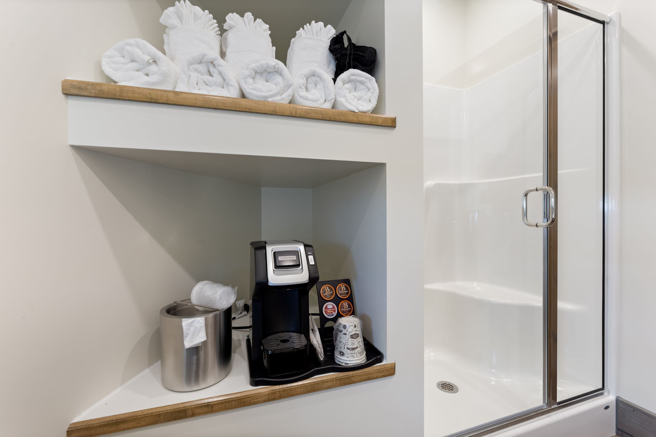 Un baño de hotel con ducha, un perchero con toallas, una cafetera y un cubo de basura.