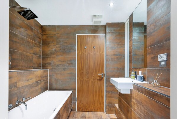 A modern bathroom with a wooden door, a white bathtub, and a sink with a mirror above it.