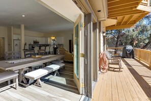 Et rummeligt spiseområde indendørs med et langt bord og bænke, et køkken i baggrunden samt en træterrasse med udendørs møbler.