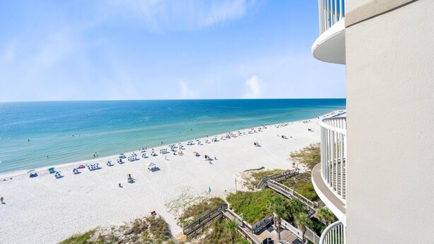 Ein Strand mit blauem Himmel, ein weißes Gebäude mit Balkon und eine Holzstege, der zum Strand führt.