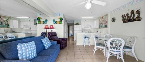 A living room with a blue sofa, white chairs, a ceiling fan, and a kitchen area with white cabinets and a microwave.